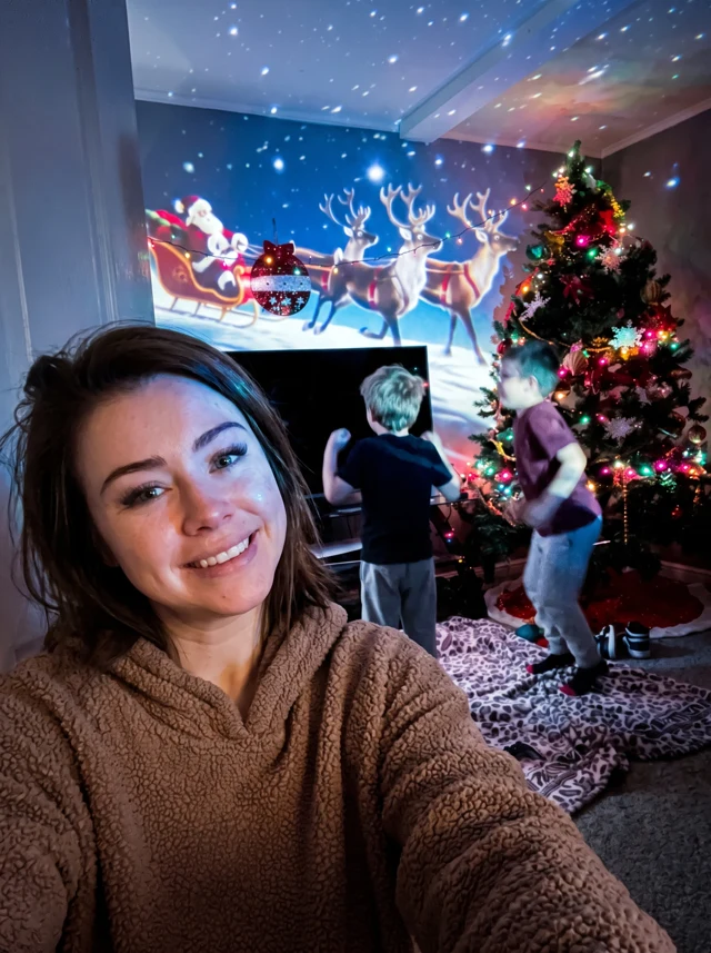 A mum taking a selfie while two children watch Santa and his reindeer projected across their Christmas living room